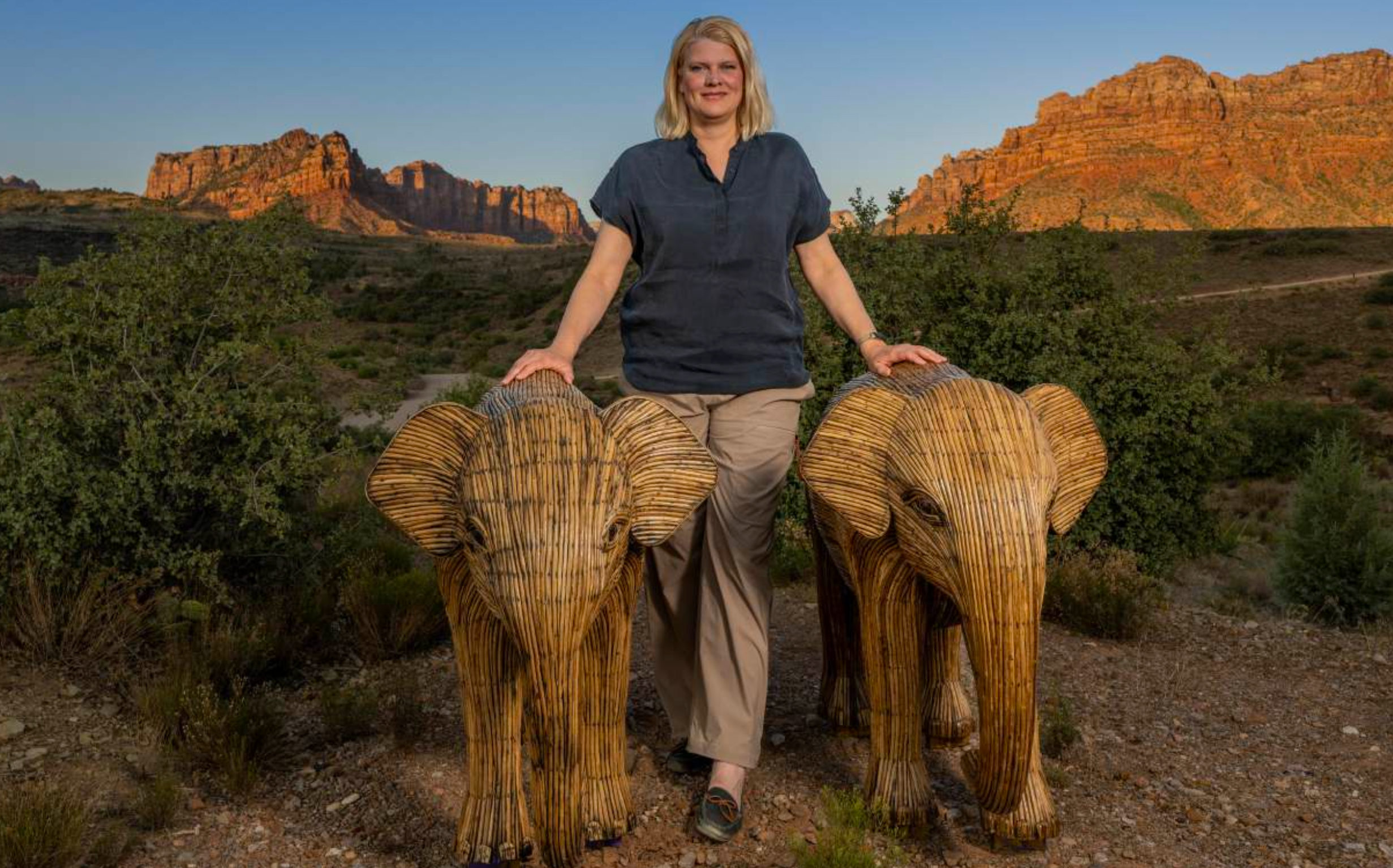 A woman stands outdoors between two large woven straw elephant sculptures in a desert landscape with rock formations and shrubs under a blue sky.